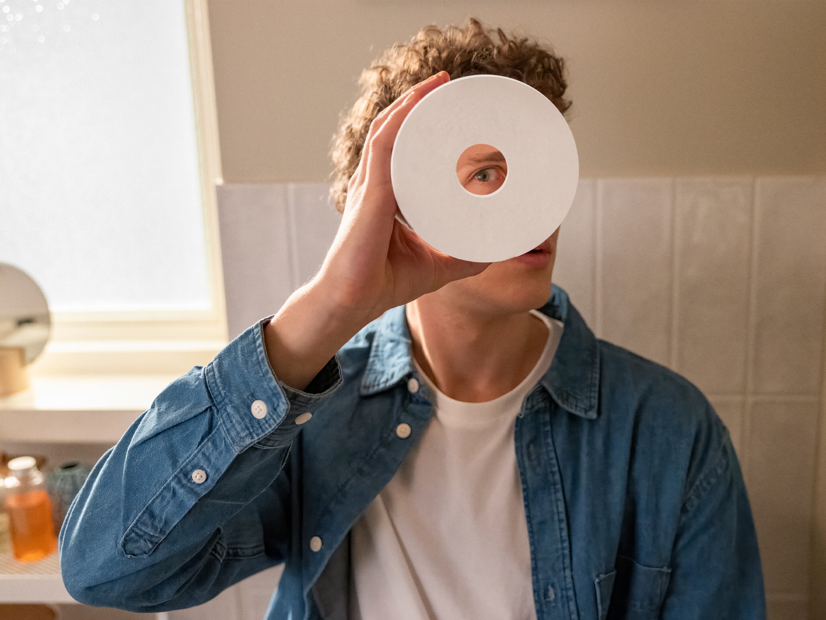 En mand i denimskjorte kigger legende gennem en toiletrulle i et lyst og moderne badeværelse.