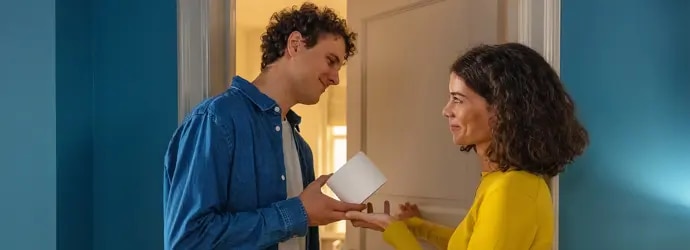 Un homme et une femme sont devant la porte d'une salle de bain, il lui tend un rouleau de papier toilette.