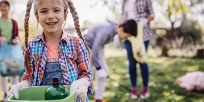 Une jeune fille tient une poubelle verte, sa famille nettoie un espace vert en arrière-plan.