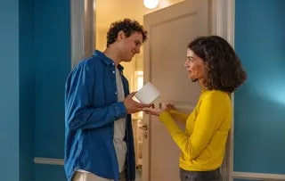 Un homme et une femme sont devant la porte d'une salle de bain, il lui tend un rouleau de papier toilette.