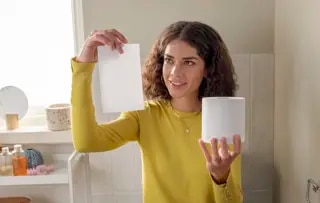 Une femme, assise dans une salle de bain, tient une feuille de papier toilette d'une main et un rouleau de l'autre.