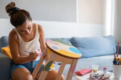 Une femme est assise à une table basse et peint un tabouret en bois à pois de différentes couleurs.