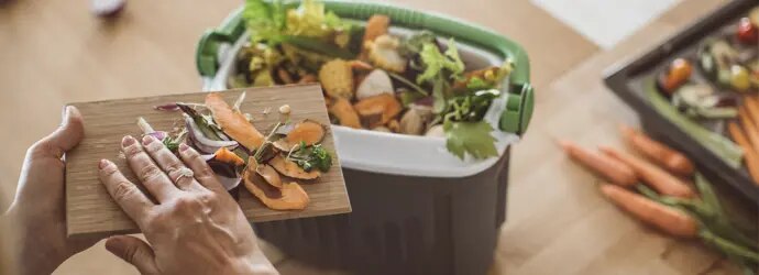 A woman scrapes vegetable scraps off of a chopping board, into a compost bin.