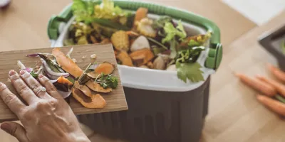 A woman scrapes vegetable scraps off of a chopping board, into a compost bin.