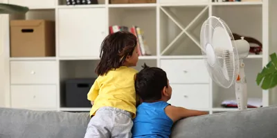 Deux enfants regardent un ventilateur depuis un canapé gris, dos contre dos.