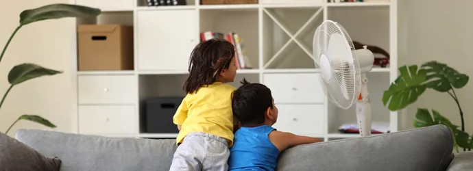 Deux enfants regardent un ventilateur depuis un canapé gris, dos contre dos.