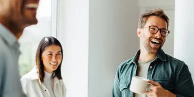 Trois personnes, dont l'une tient une tasse à café, sourient dans une pièce lumineuse.