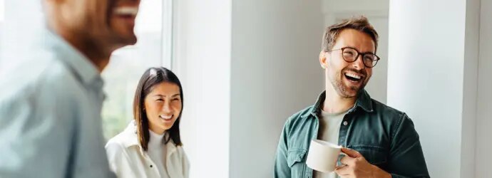 Trois personnes, dont l'une tient une tasse à café, sourient dans une pièce lumineuse.