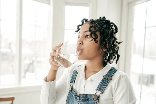 Une jeune personne se tient dans une pièce très éclairée et boit un verre d'eau.