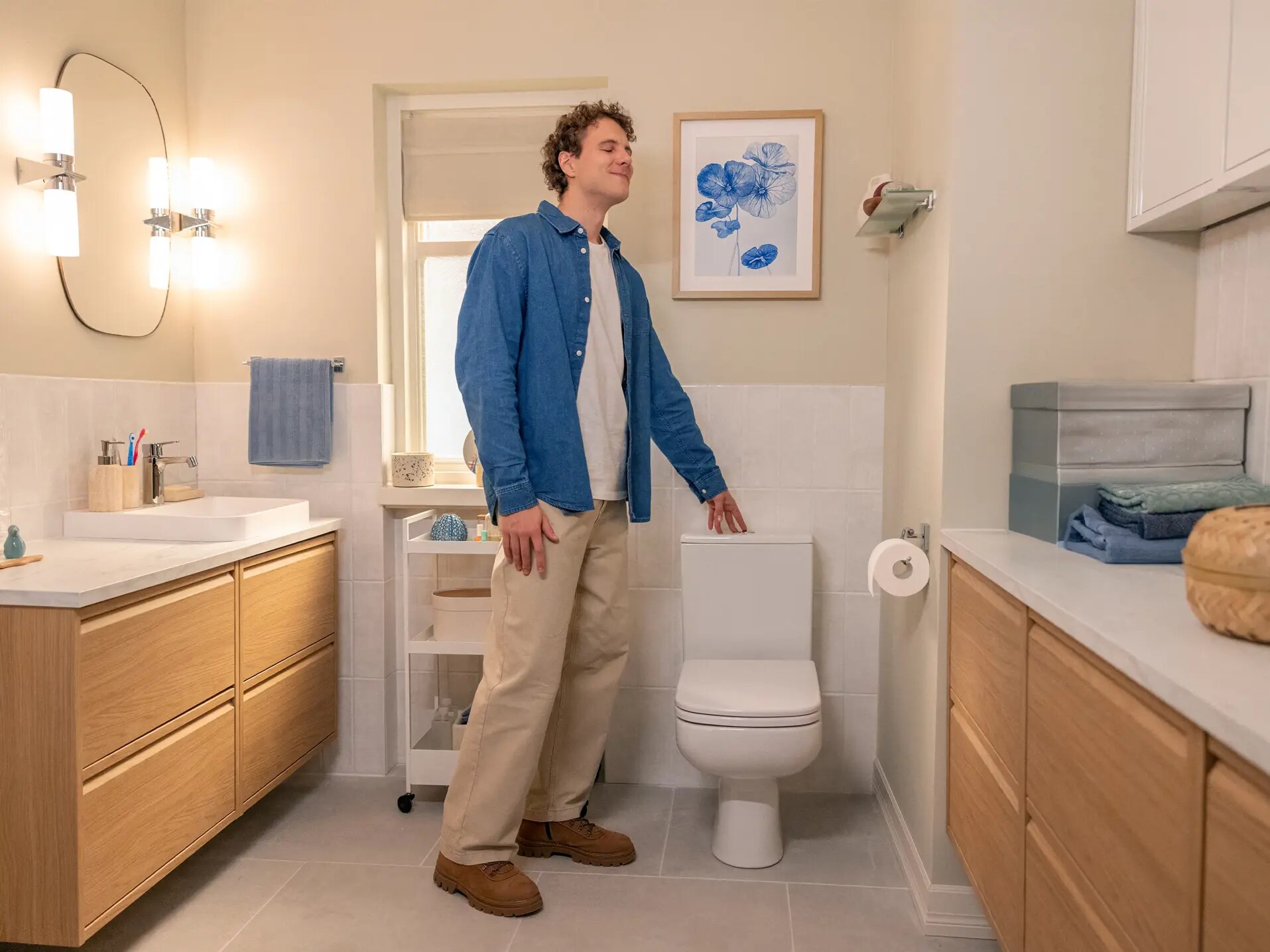 Mies hymyilee modernissa kylpyhuoneessa wc:n ja puukaappien vieressä valoisassa ja siististi suunnitellussa tilassa.